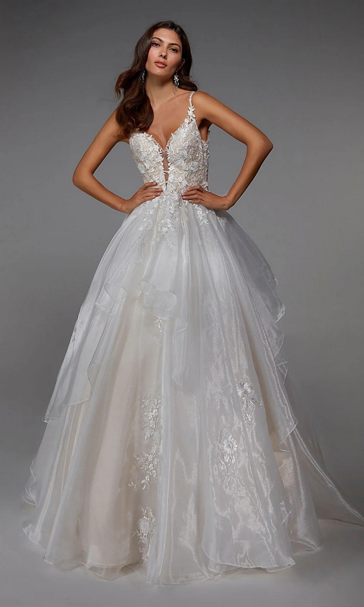 A woman stands confidently with hands on hips, wearing the Alyce 7047 long white dress in ivory/sand, featuring a sleeveless, floor-length silhouette, deep V-neckline, lace bodice, and layered tulle skirt.